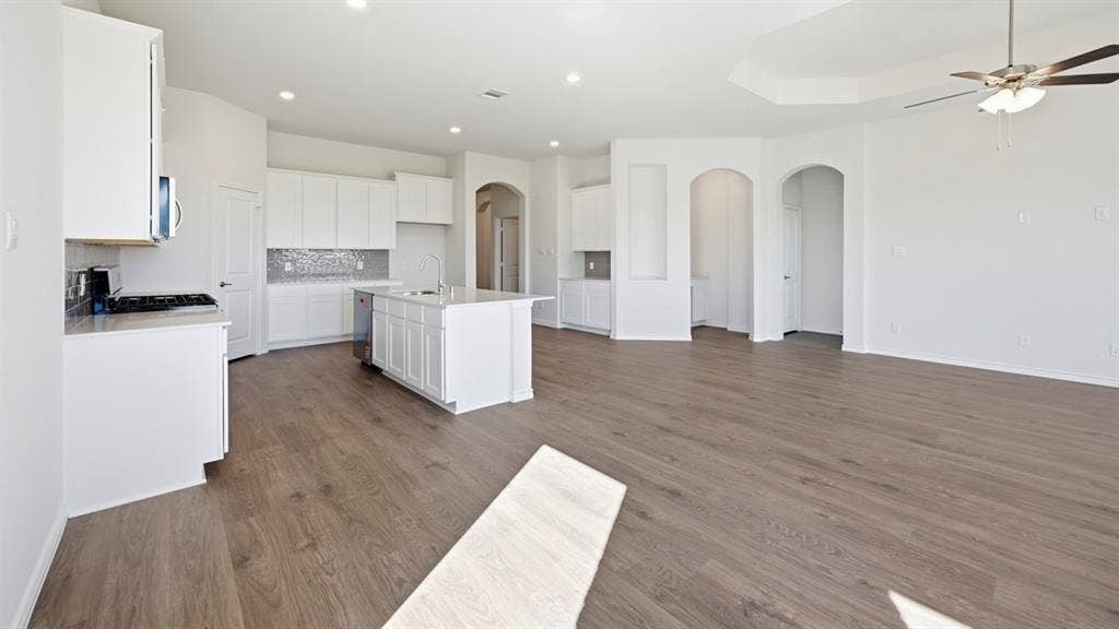 2506 Wesley Avenue, Royse City, TX 75189 Open-concept interior featuring light wood-finish flooring, a contemporary kitchen with white cabinetry and stainless steel appliances, a center island with sink, a decorative tile backsplash, and a spacious living area with a tray ceiling and ceiling fan