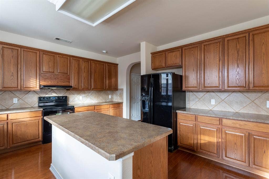 6509 Hickory Hill Drive, Plano, TX 75074 Kitchen featuring wood-finish cabinetry, a central island with a solid surface countertop, tile backsplash, and black appliances