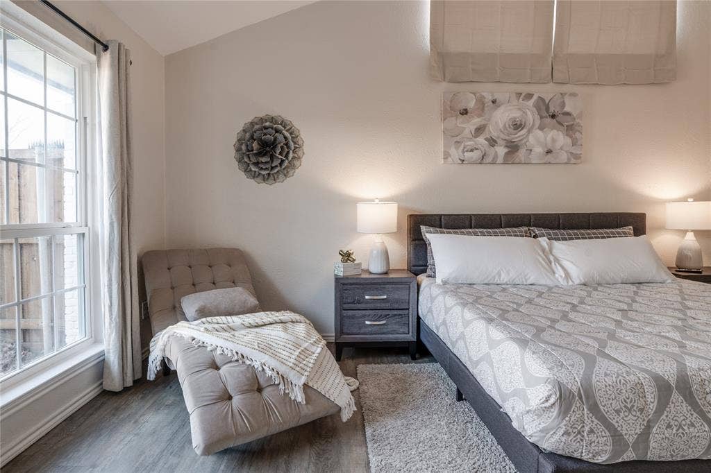 Vaulted ceiling, large window with grid pattern, wood-finish flooring, paneled headboard, and dual nightstands with integrated lighting