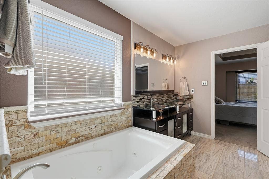1125 Bayfield Drive, Denton, TX 76209 Jetted soaking tub with stone tile surround, dual vanity with dark wood finish cabinetry, mosaic tile backsplash, and a large mirror with overhead lighting