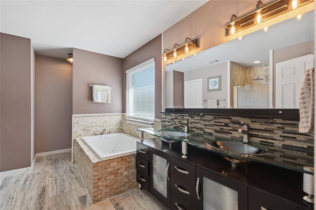 1125 Bayfield Drive, Denton, TX 76209 Dual vessel sinks, stone tile backsplash, dark wood-finish vanity, built-in jetted tub, and large format tile flooring