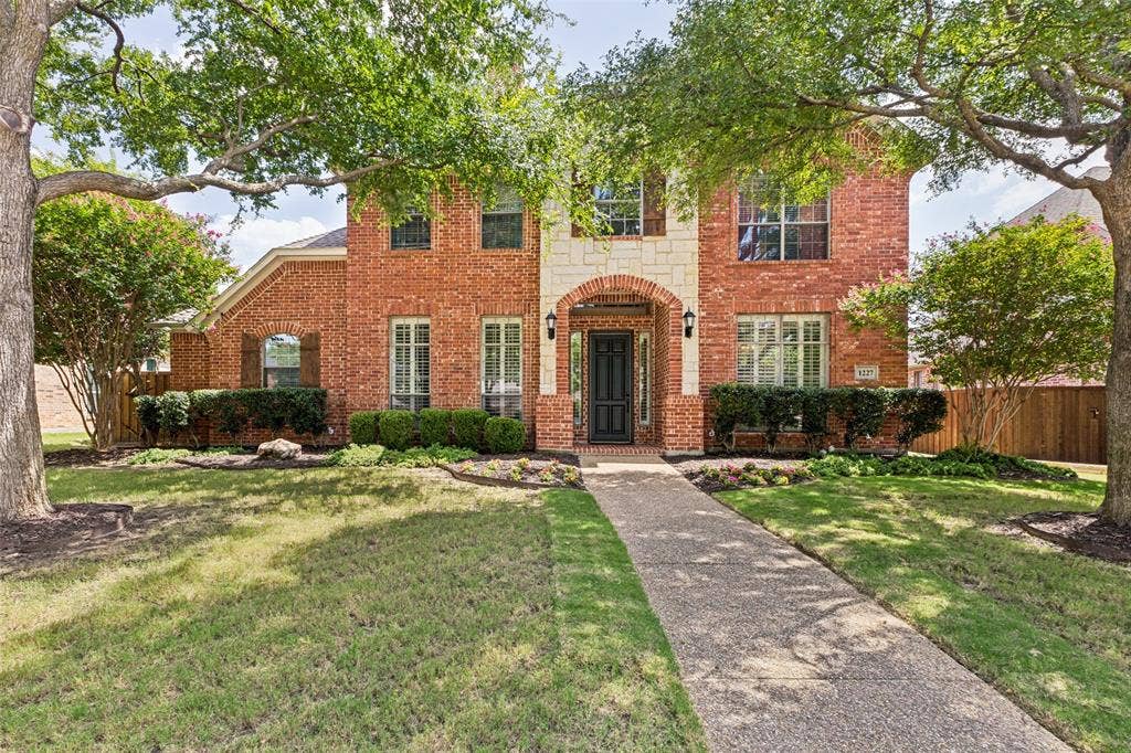1227 Comanche Drive, Allen, TX 75013 Brick and stone exterior featuring an arched entry, a dark paneled door, multiple window styles, and mature trees framing the property
