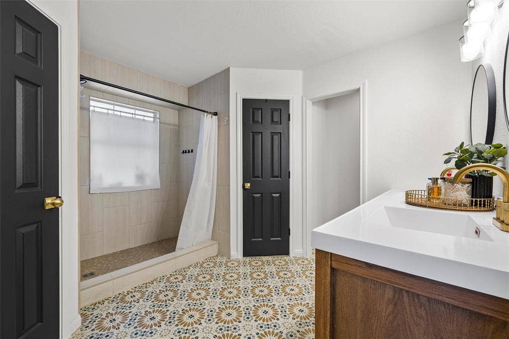 Patterned tile flooring, walk-in shower with tiled walls, dark paneled doors, white countertop, and wood-finish vanity with an integrated sink