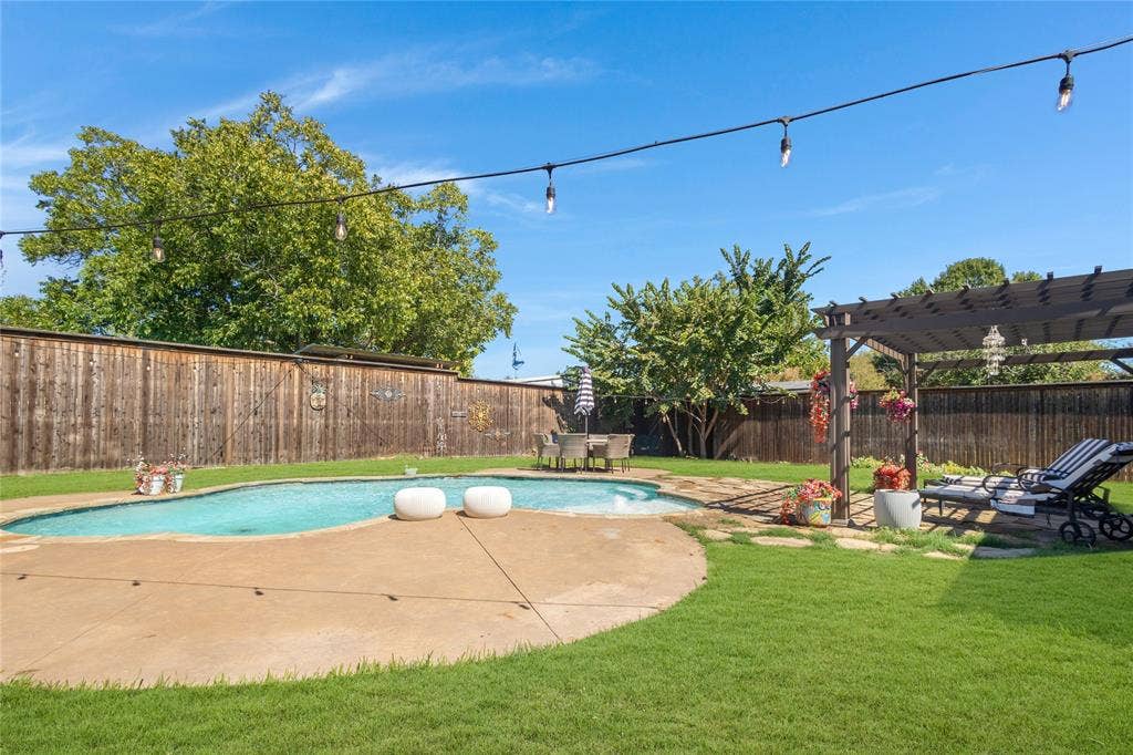 Expansive backyard featuring a curved swimming pool, concrete patio, lush lawn, wood privacy fencing, and a built-in pergola