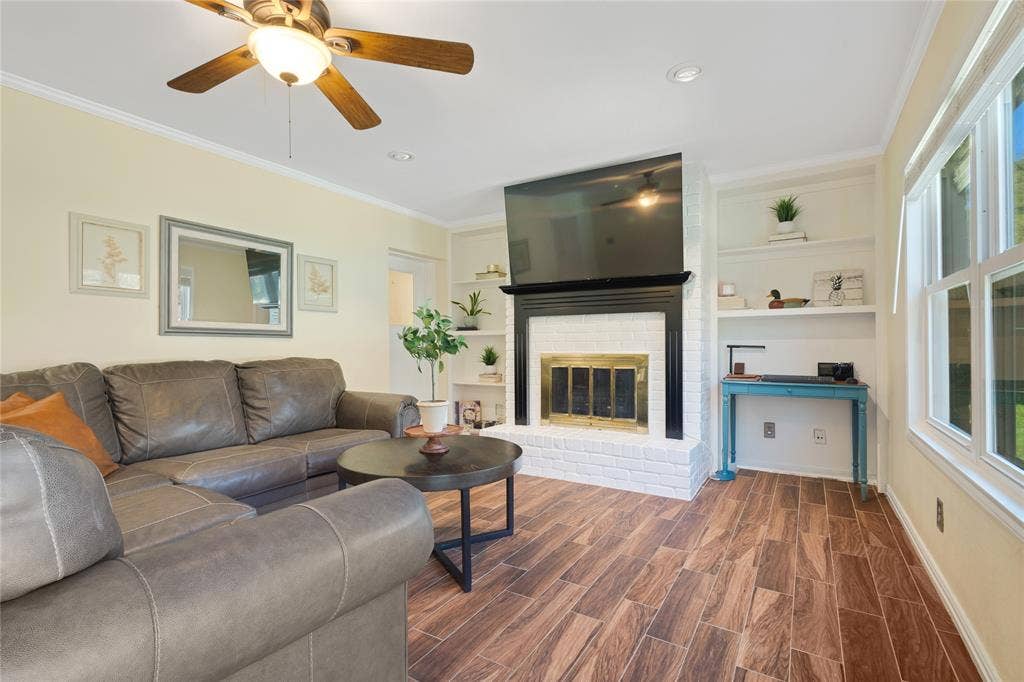 Wood-finish flooring, white brick fireplace with a black wood mantel, built-in shelving, multi-pane windows, and a ceiling fan
