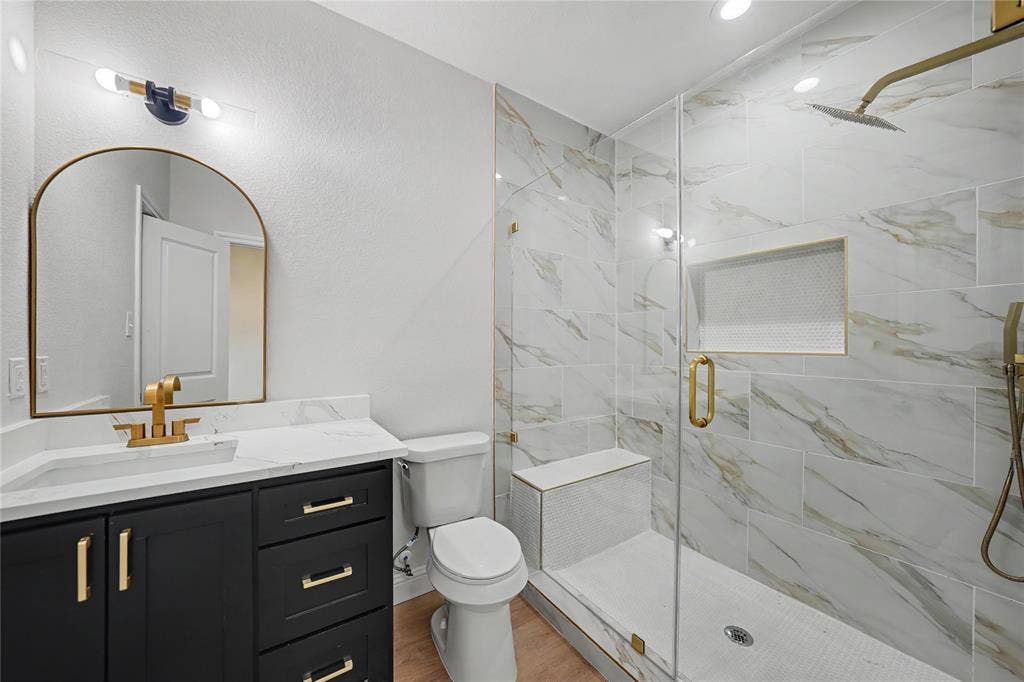 817 Stamps Avenue, Fort Worth, TX 76114 Modern bathroom featuring a black vanity with gold hardware, a white countertop, an arched gold-framed mirror, and a glass-enclosed shower with white and gold-veined tile