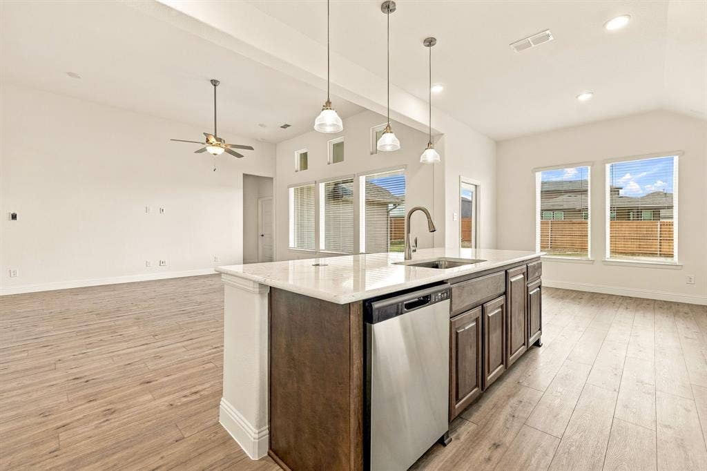 Kitchen island featuring stone countertops, integrated stainless steel dishwasher, undermount sink with gooseneck faucet, and dark-toned cabinetry, complemented by wood-finish flooring, vaulted ceilings, and pendant lighting