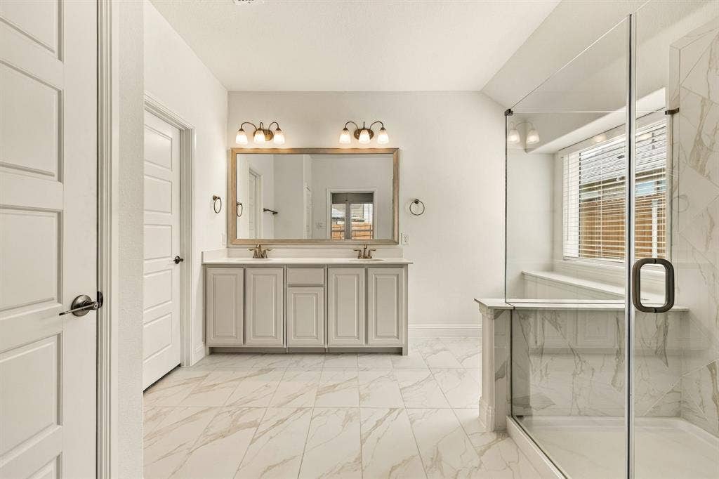 Dual vanity with light-toned cabinetry and solid surface countertop, expansive framed vanity mirror, glass-enclosed shower with stone-finish tile surround, and neutral stone-finish floor tiling