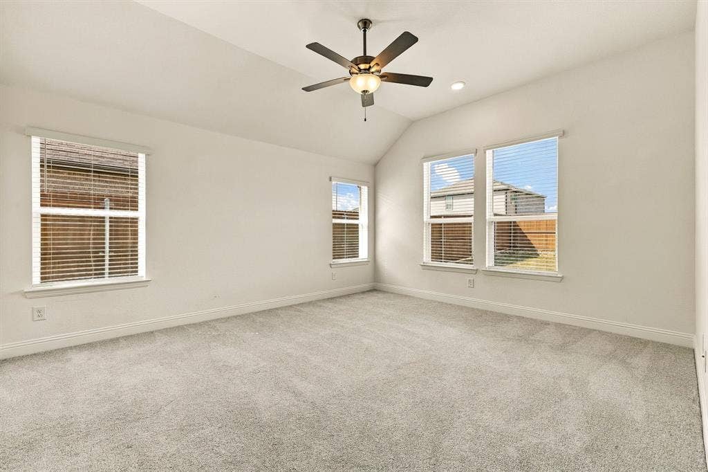 Vaulted ceiling featuring a central five-blade ceiling fan with integrated lighting, neutral wall-to-wall carpeting, and multiple windows with horizontal blinds