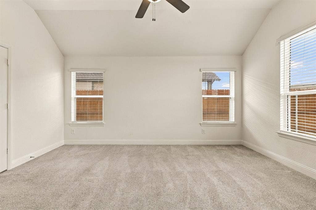 Vaulted ceiling with a centrally mounted ceiling fan, neutral wall-to-wall carpeting, and multiple windows with installed horizontal blinds