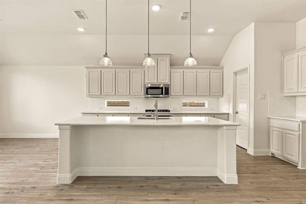 Kitchen featuring a central island with stone countertop, wood-finish flooring, white shaker-style cabinetry, a herringbone-patterned backsplash, and trio of pendant lights