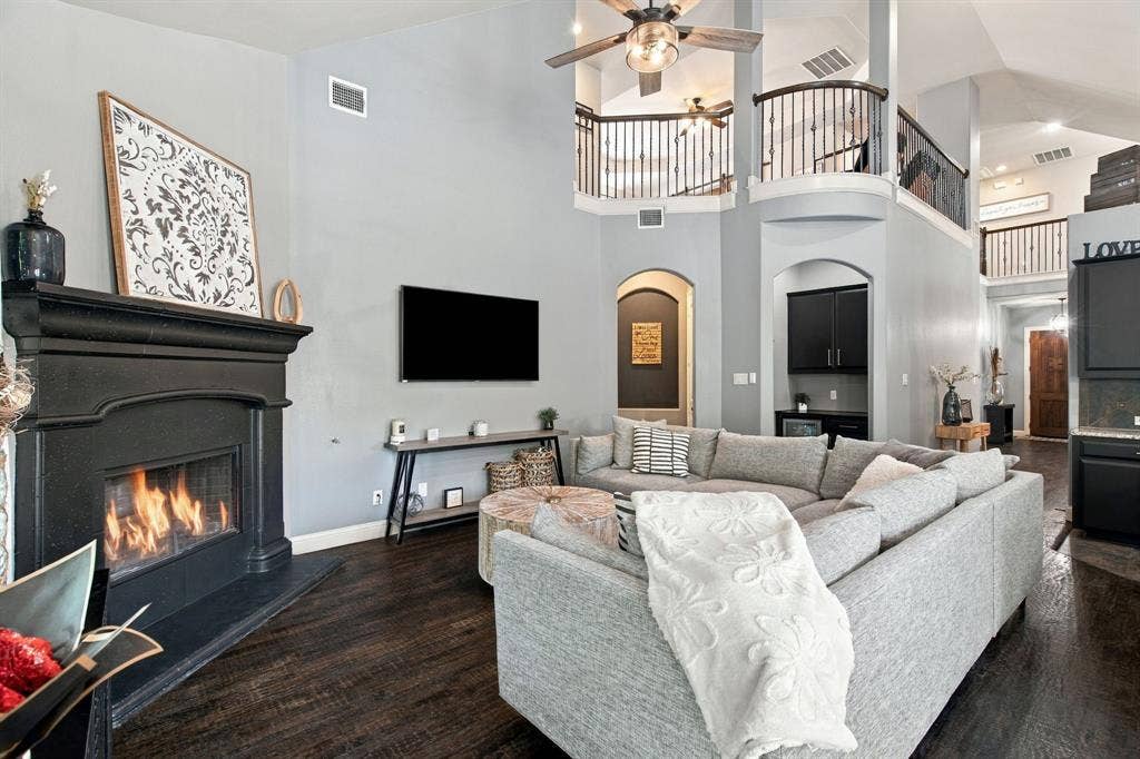 Spacious living area centered around a statement fireplace with a dark mantel, complemented by soaring ceilings and an open view of the upstairs balcony.