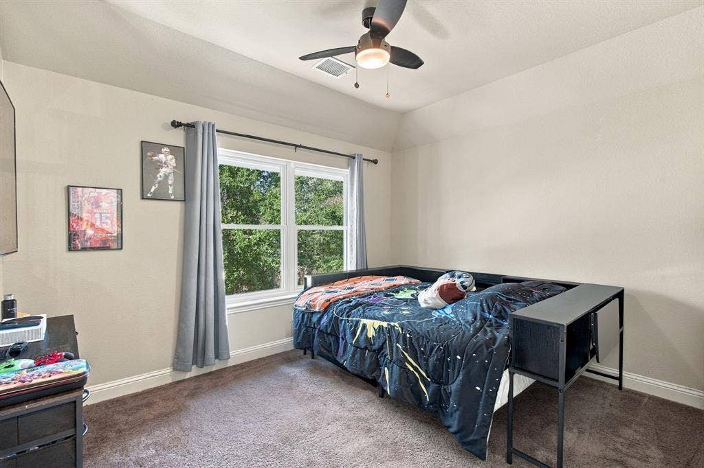 Comfortable upstairs bedroom featuring a large window with leafy views, ceiling fan, neutral walls, and carpeting.