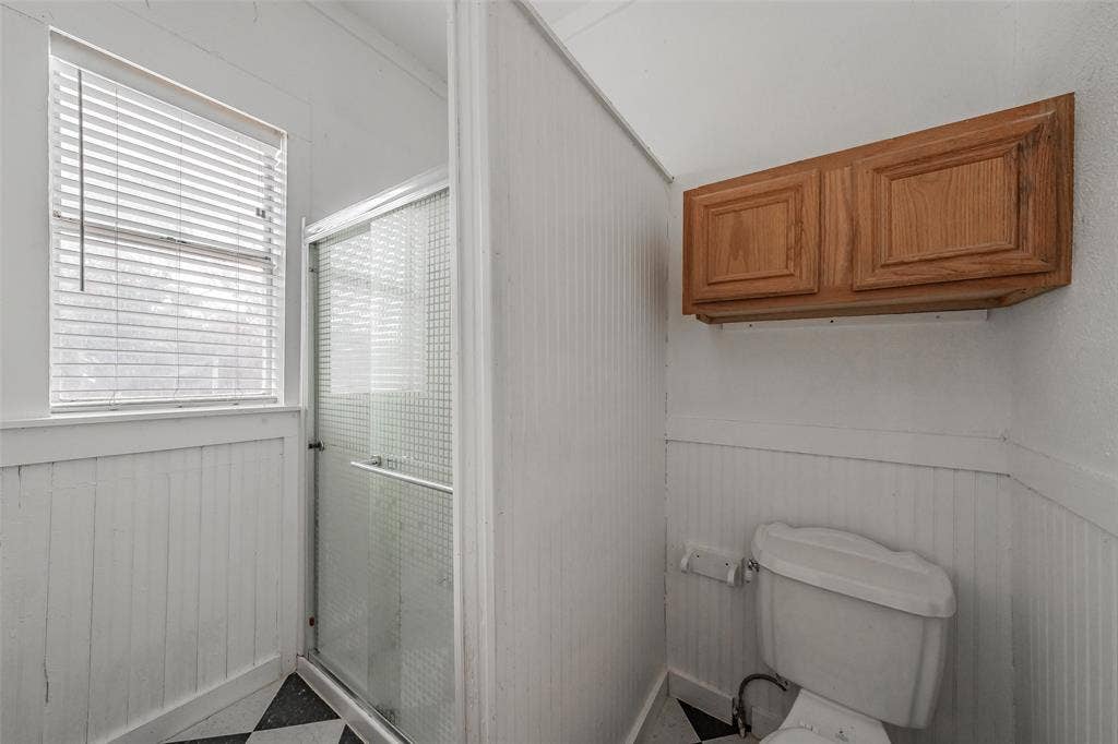 212 N Ruddell Street, Denton, TX 76205 Shower enclosure with glass door, white beadboard paneling, black and white checkered flooring, and natural wood finish wall-mounted cabinetry