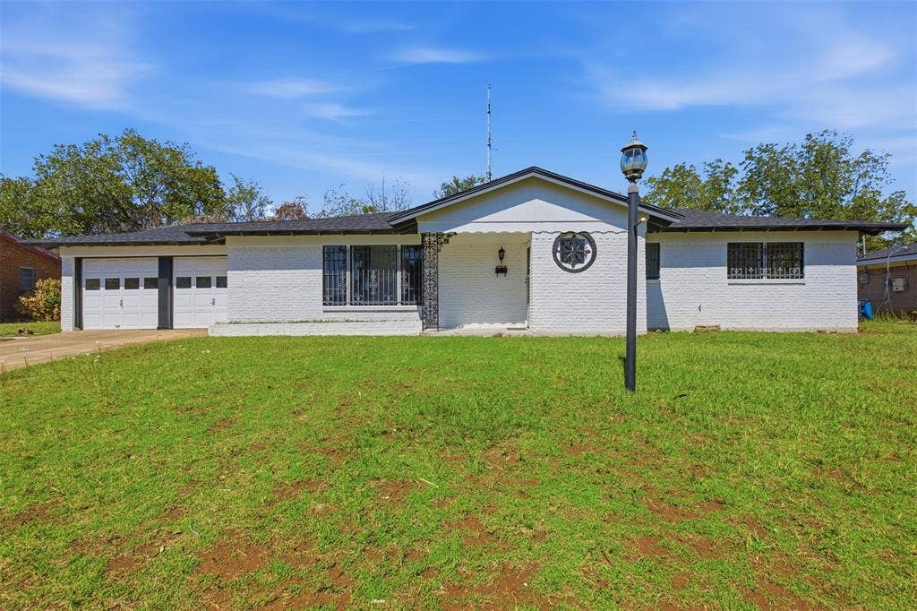2313 Clearview Drive, Fort Worth, TX 76119 White brick exterior with a black shingle roof, featuring a two-car garage with white paneled doors, and a decorative wrought iron entry