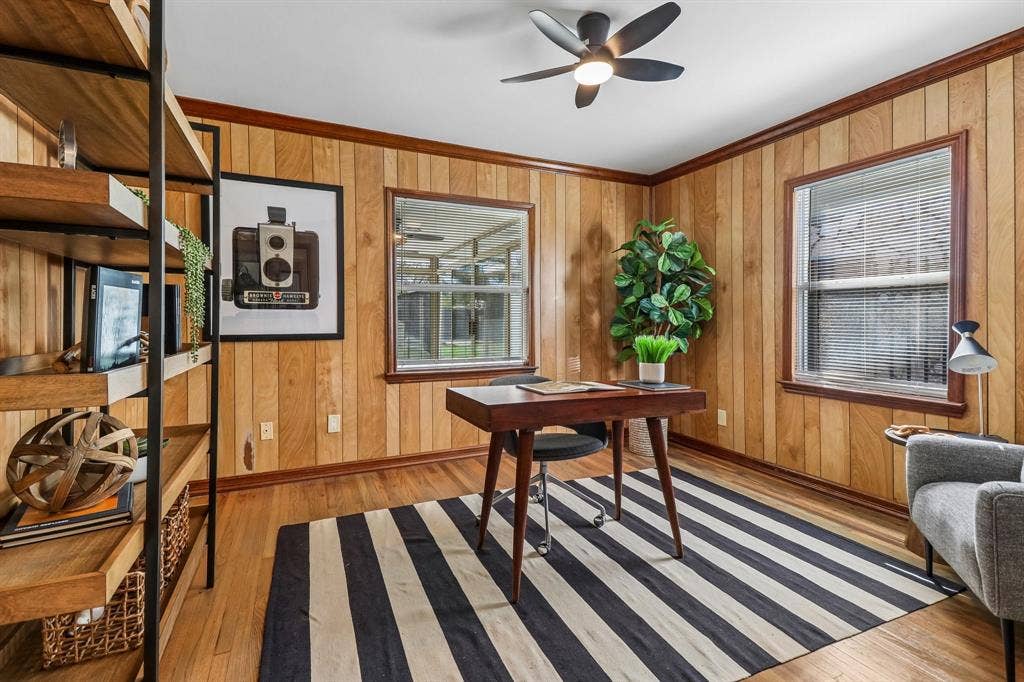 Wood-paneled walls with wood trim, wood-finish flooring, two windows with blinds, and a ceiling fan with integrated lighting