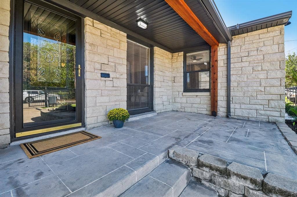 Stone veneer facade, glass-paneled entry door, dark-framed windows, wood beam accents, and a stone patio with integrated steps