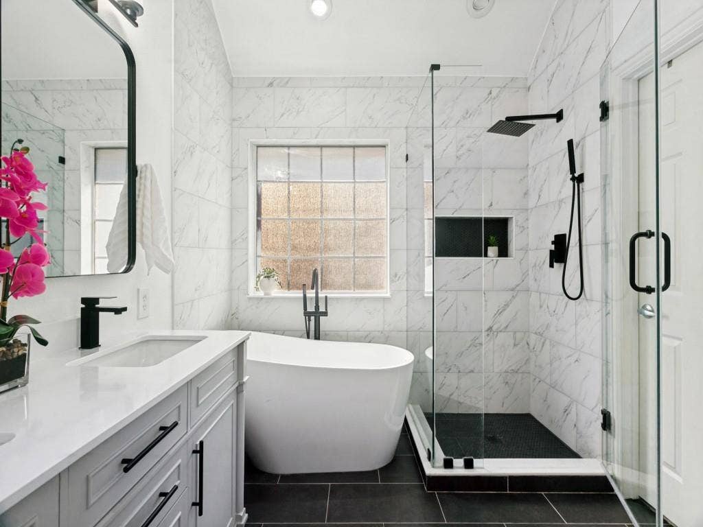 Freestanding soaking tub, glass-enclosed shower with black fixtures, vanity with quartz countertop, and large-format wall tiling