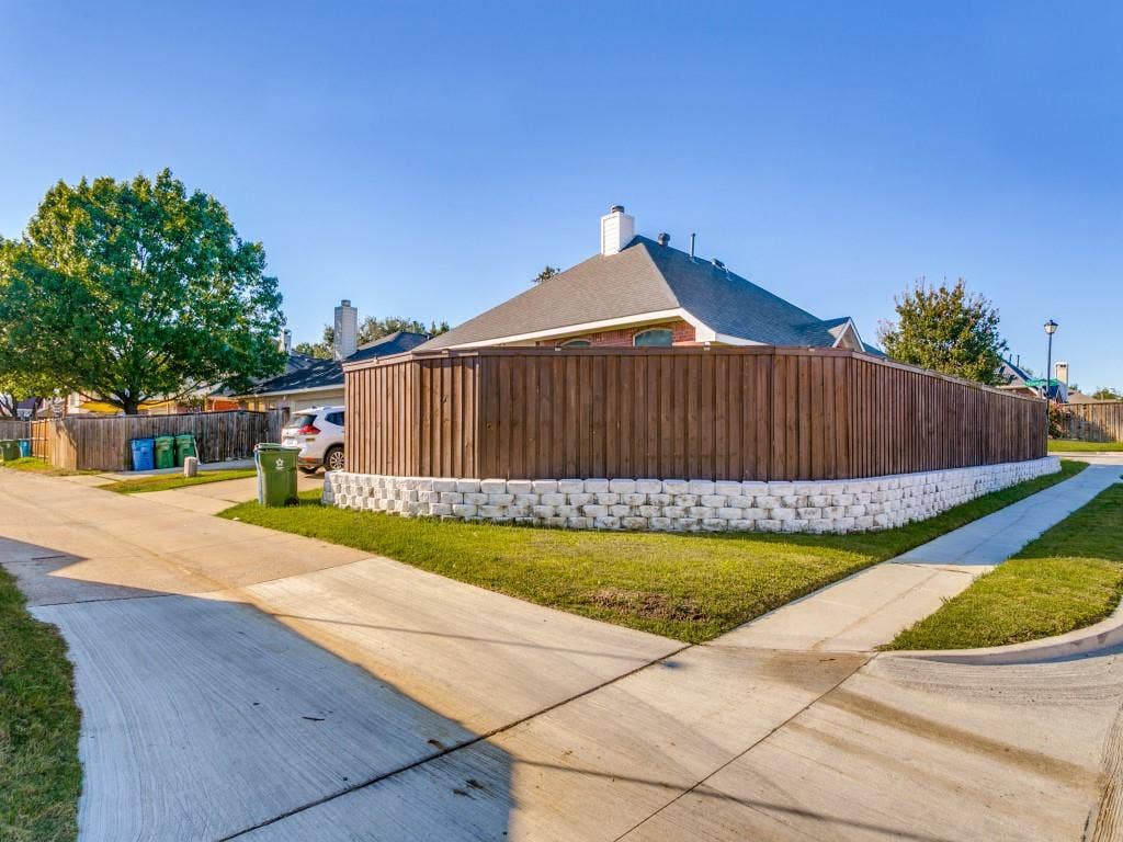 Wood-finish privacy fence with a white retaining wall, asphalt shingle roof, and mature perimeter trees