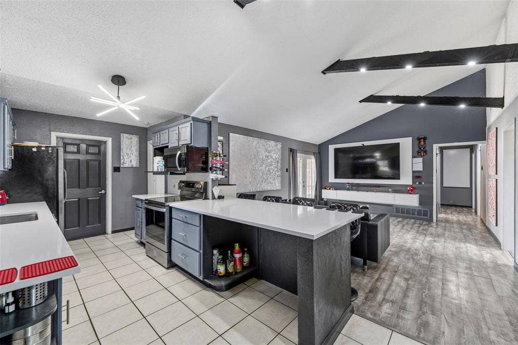 6606 Coleshire Drive, Dallas, TX 75232 Vaulted ceiling with exposed beams, wood-finish flooring, white countertop island, stainless steel appliances, and ceiling-mounted light fixture