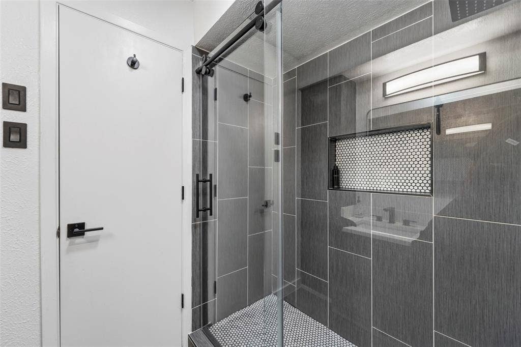 6606 Coleshire Drive, Dallas, TX 75232 Shower with dark gray rectangular tile, black penny tile flooring, built-in recessed shelf with penny tile, and sliding glass enclosure with black hardware