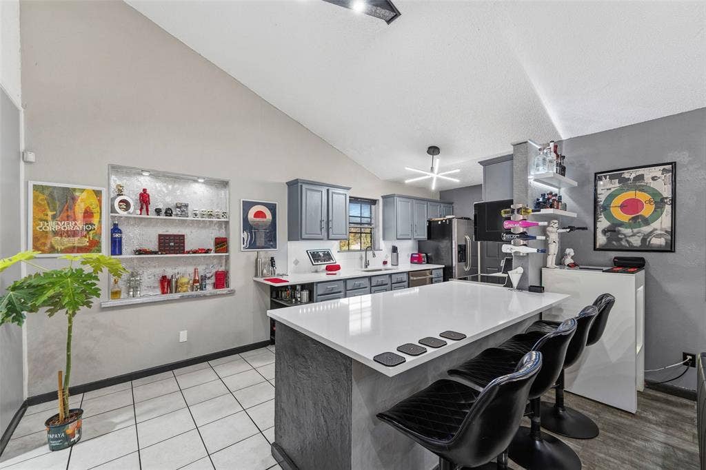 6606 Coleshire Drive, Dallas, TX 75232 Kitchen featuring a vaulted ceiling, recessed shelving with integrated lighting, a central island with countertop seating, gray cabinetry, and tiled flooring