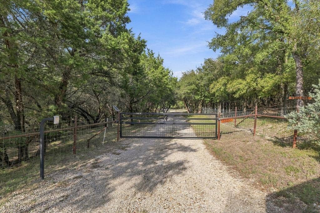 1600 Fox Ridge Road, Lorena, TX 76655 Gated entrance featuring a metal-frame drive gate, wire perimeter fencing, and a gravel driveway bordered by dense mature trees