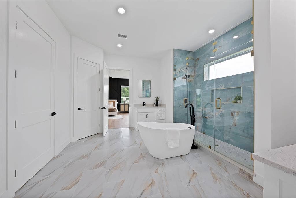 Freestanding soaking tub, walk-in shower with glass enclosure, dual vanity with quartz countertop, light-toned tile flooring, and recessed lighting