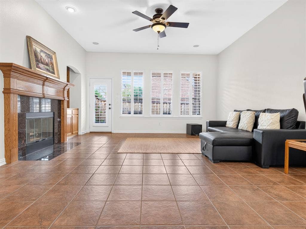 Vaulted ceilings with recessed lighting, an ornate wood-mantle fireplace with a dark stone surround, tiled flooring, a ceiling fan, and multiple windows with plantation shutters