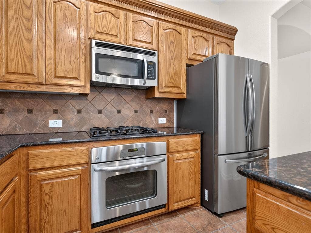 Hardwood cabinetry, stainless steel appliances, dark stone-finish countertops, ceramic tile backsplash, and ceramic tile flooring