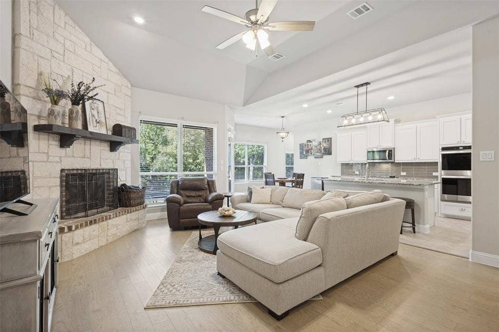 Vaulted ceiling with a ceiling fan, stone-clad fireplace with a hearth, wood-finish flooring, kitchen with white cabinetry and stainless steel appliances, and a kitchen island with seating