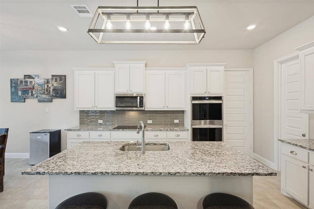 Kitchen featuring an island with stone countertop and undermount sink, white cabinetry with silver hardware, stainless steel built-in double oven, and gray tile backsplash