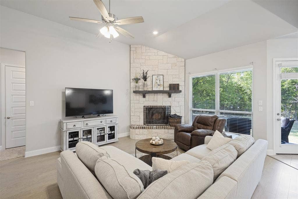 Vaulted ceiling with a ceiling fan, stone fireplace with a wood mantel, light wood-finish flooring, large windows, and a glass-paneled door