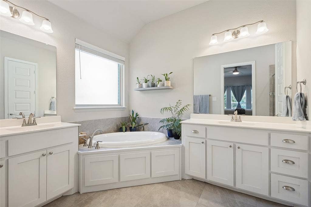 Dual vanity sinks with white cabinetry, a built-in soaking tub, a window with privacy blinds, and tile flooring