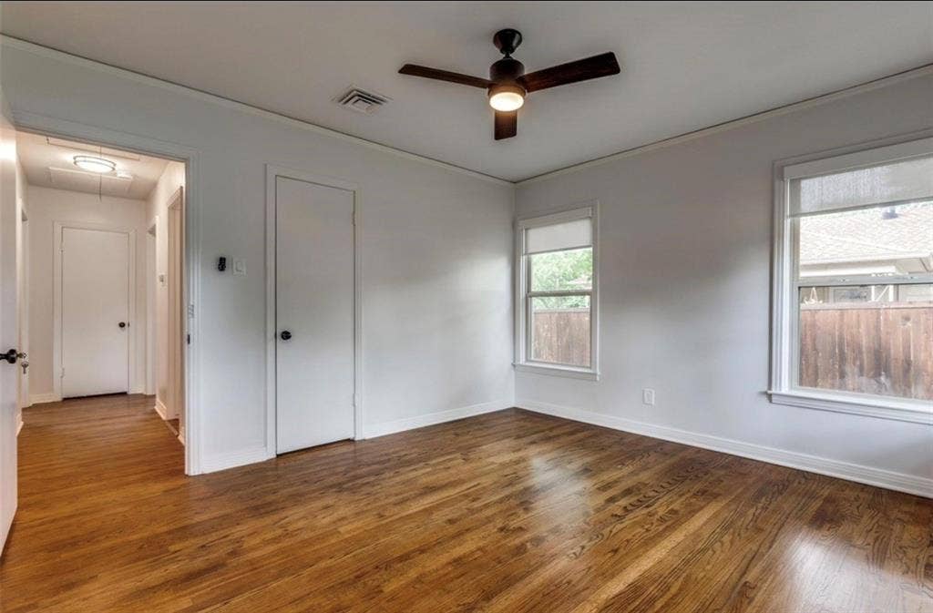 Rich wood-finish flooring, two windows with roller shades, a ceiling fan with integrated lighting, and a white interior door