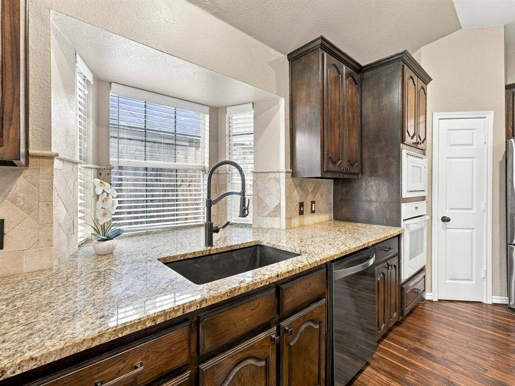 Kitchen featuring wood-finish flooring, granite countertops, dark wood cabinetry, a built-in oven, and a large sink with a gooseneck faucet