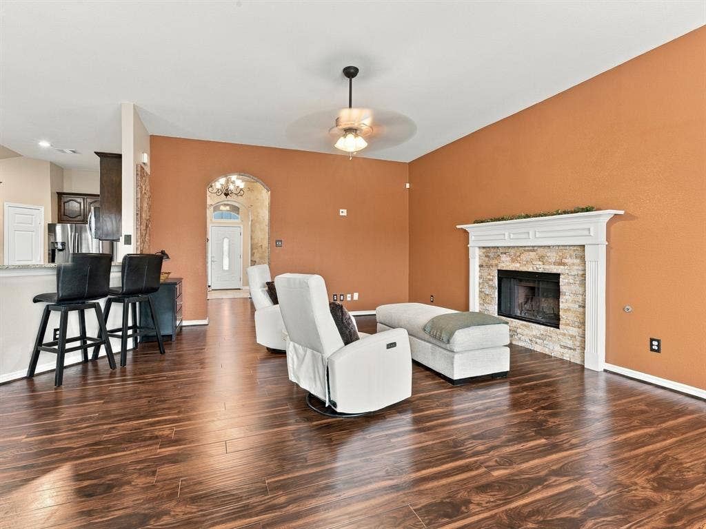 Spacious living area featuring wood-finish flooring, a stacked stone fireplace with a white mantel, and an arched entryway