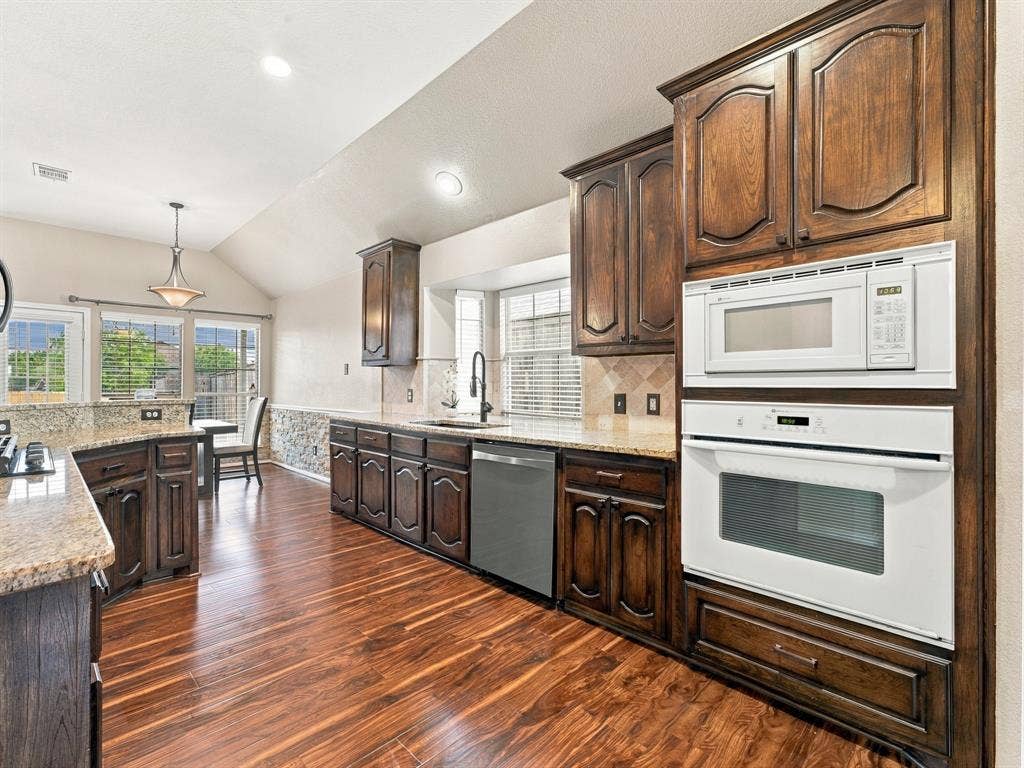 Vaulted ceiling with recessed lighting, wood-finish flooring, built-in oven and microwave, dark wood cabinetry, and light-toned countertops