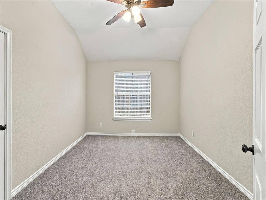 Vaulted ceiling, ceiling fan with light fixture, window with blinds, light gray carpet, and white trim molding