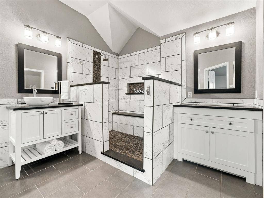 Walk-in shower with tiled walls and floor, dual vanities with vessel sinks, and grey tile flooring