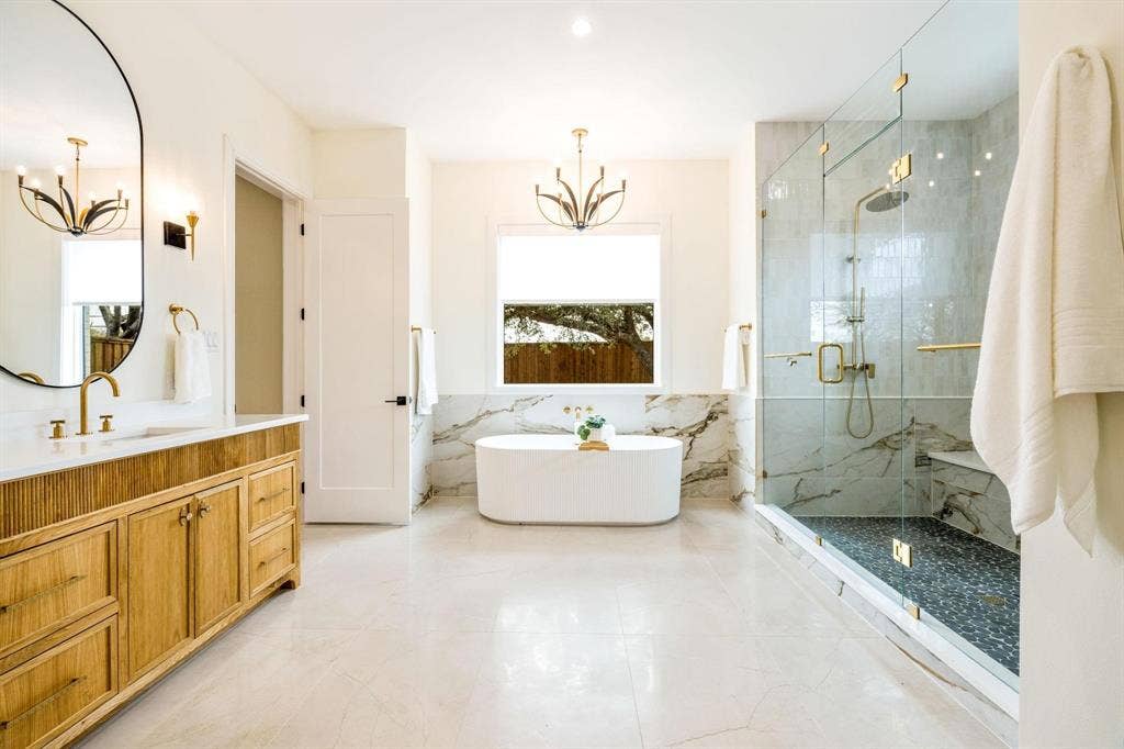 12007 Quincy Lane, Dallas, TX 75230 Freestanding oval bathtub, glass-enclosed shower with a built-in bench, wood-finish vanity with a stone countertop, and an oval wall mirror