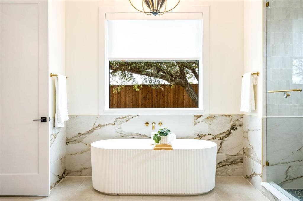 12007 Quincy Lane, Dallas, TX 75230 Freestanding soaking tub with a wall-mounted faucet, complemented by large format tile with natural veining, and a glass-enclosed shower with brass hardware