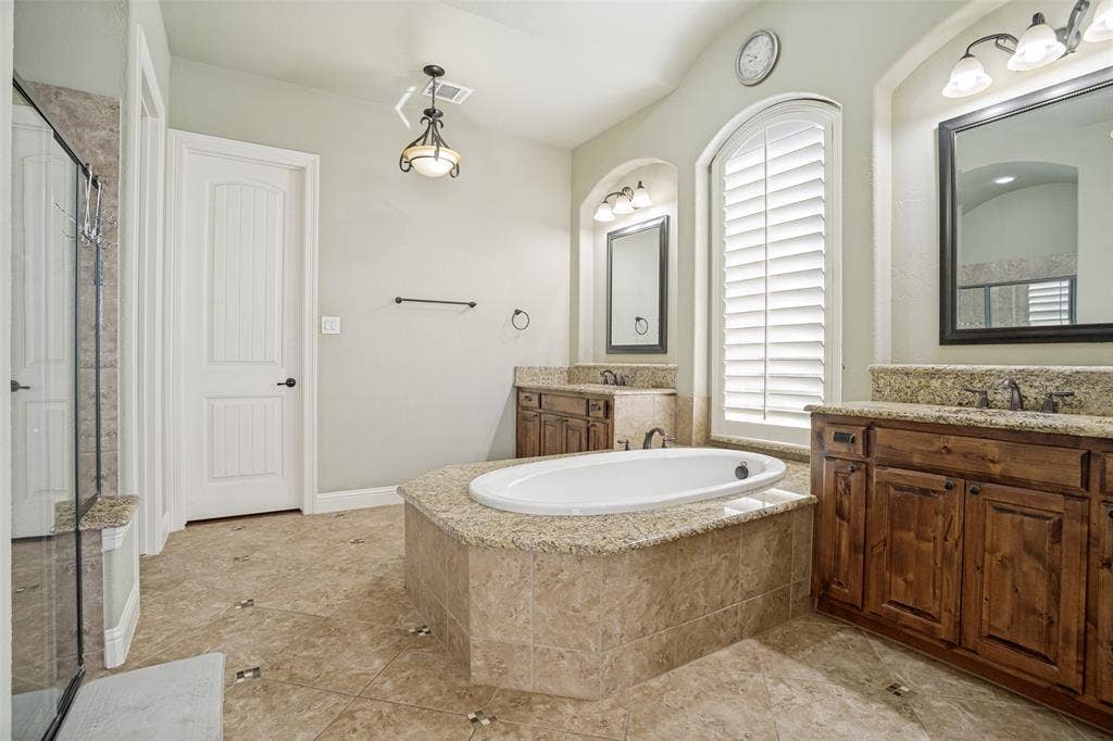 1250 Packsaddle Trail, Prosper, TX 75078 Spacious bathroom featuring a built-in soaking tub with a stone surround, dual wood vanities with stone countertops, and an arched window with plantation shutters