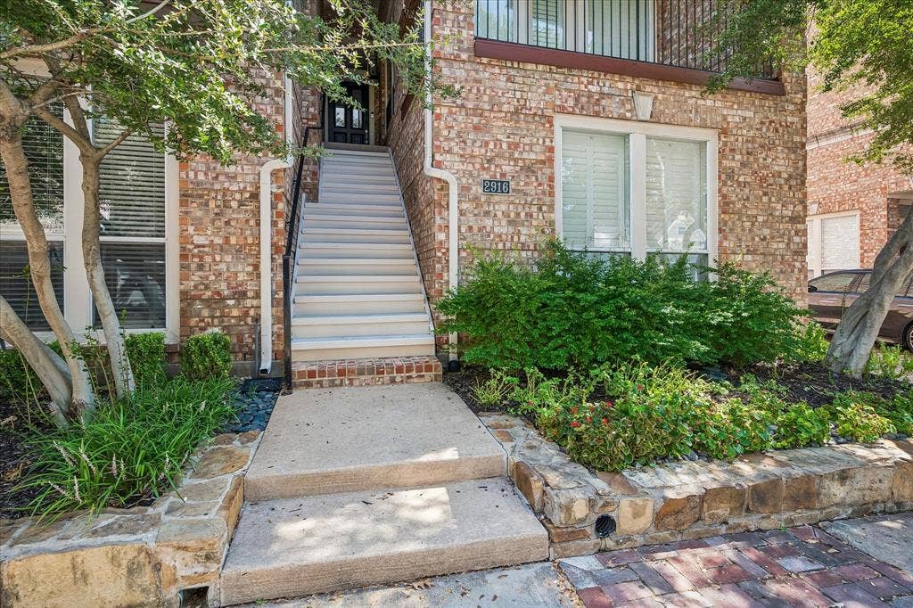 2916 Thomas Avenue, Dallas, TX 75204 Brick exterior with white window frames, a stone retaining wall, and a concrete pathway leading to an exterior staircase with dark metal railings