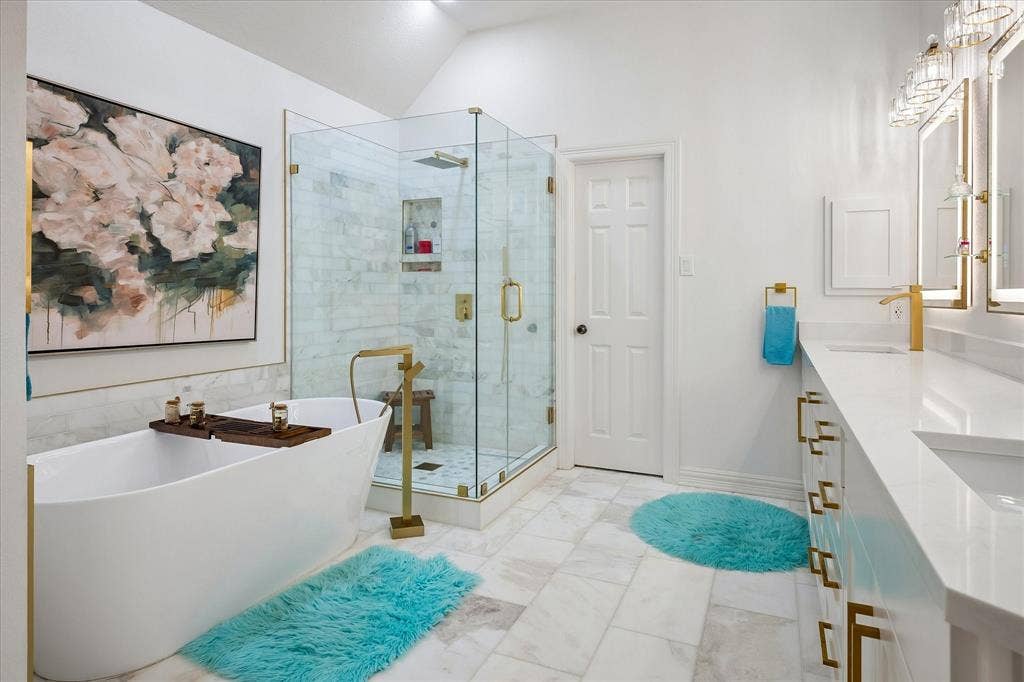 2916 Thomas Avenue, Dallas, TX 75204 Spa-inspired primary bathroom featuring a freestanding soaking tub, walk-in shower with glass enclosure and brass fixtures, dual vanities with white countertops, polished tile flooring, and framed floral artwork