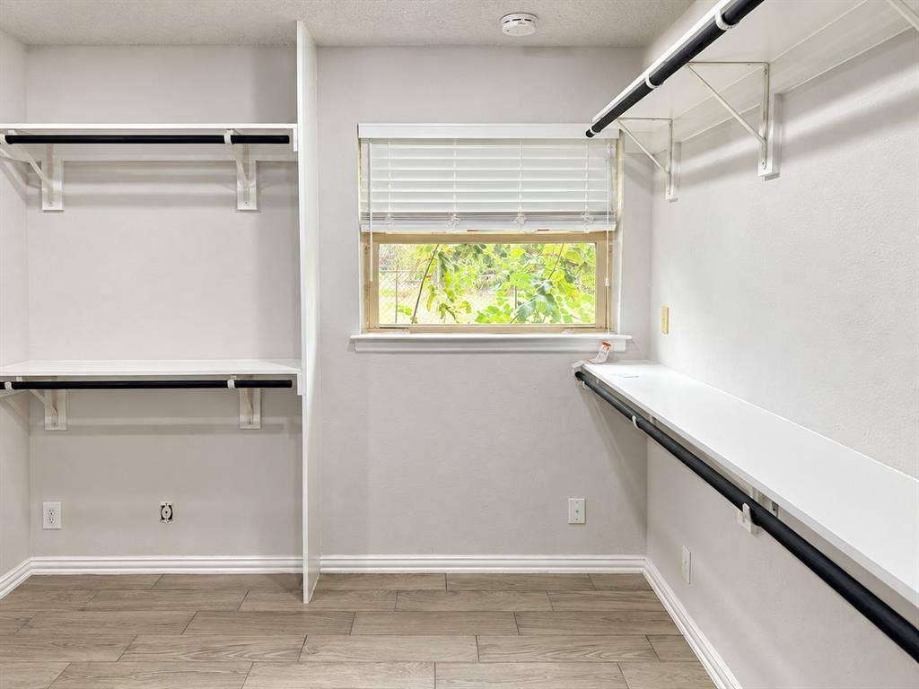 Walk-in storage space featuring wood-finish flooring, white built-in shelving, black hanging rods, and a window with horizontal blinds