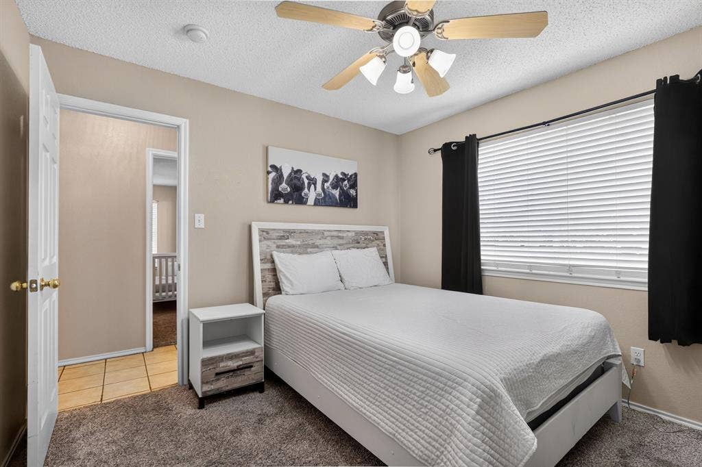 Neutral wall color, ceiling fan with light fixture, window with blinds, carpeted flooring, and a white door with brass hardware