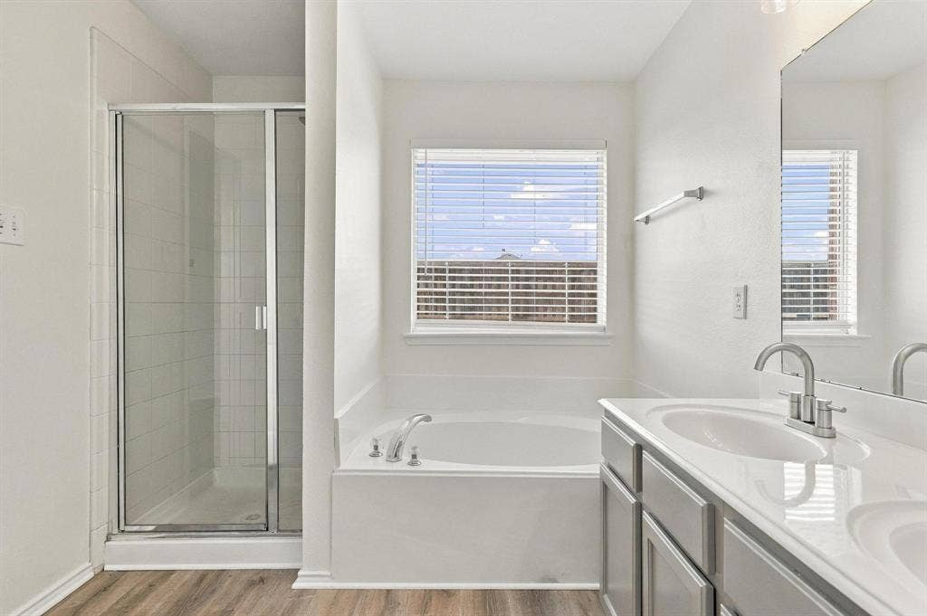 Dual vanity with undermount sinks, wood-finish flooring, a built-in tub, a separate glass-enclosed shower, and a large window