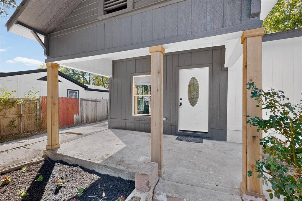 651 Ezekial Avenue, Dallas, TX 75217 Covered porch with natural wood columns, gray vertical siding, a white entry door with oval glass insert, and a landscaped planting area