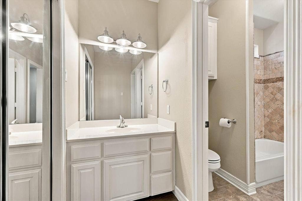 White vanity with integrated sink and chrome faucet, large vanity mirror with a four-light fixture, tile flooring, and a bath with patterned tile surround
